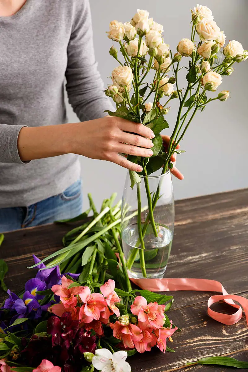 Una imagen vertical de la mano de una mujer desde la izquierda del marco dispone rosas blancas en un jarrón de vidrio colocado sobre una superficie de madera.  A la izquierda del jarrón hay flores rojas y moradas.