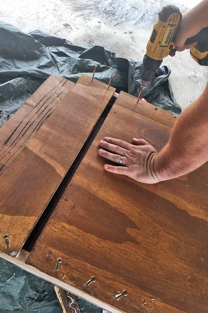 Primer plano de un hombre perforando tornillos en una caja de madera teñida de marrón con una mano mientras la mantiene firme con la otra, sobre una lona negra en la parte superior de un piso de cemento.
