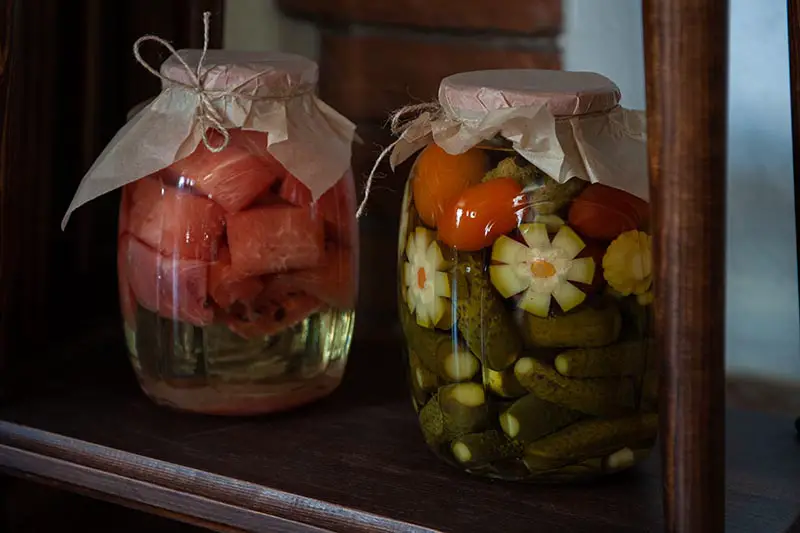 Una imagen horizontal de primer plano de dos frascos de vidrio que contienen verduras y frutas enlatadas en un estante de madera.