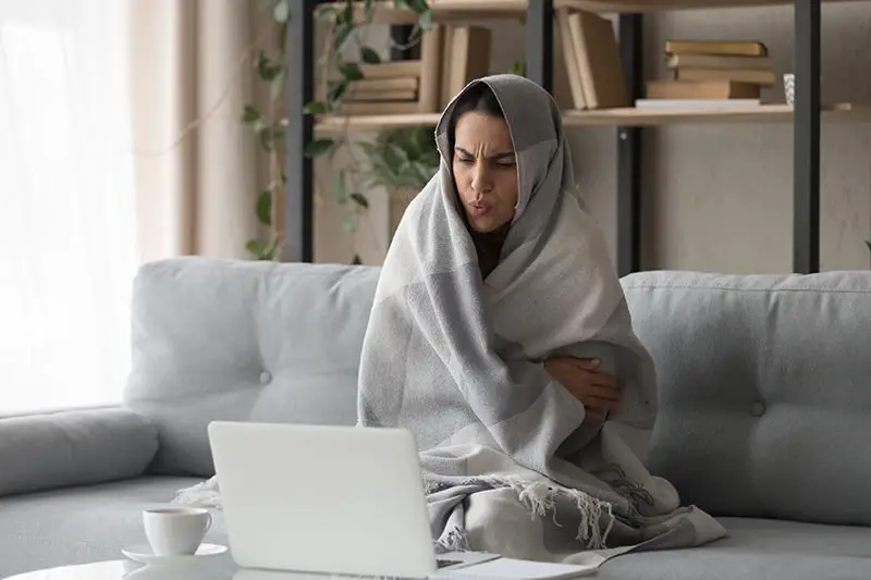Una imagen horizontal de una mujer sentada en el sofá envuelta en una manta mirando una computadora portátil durante el invierno.