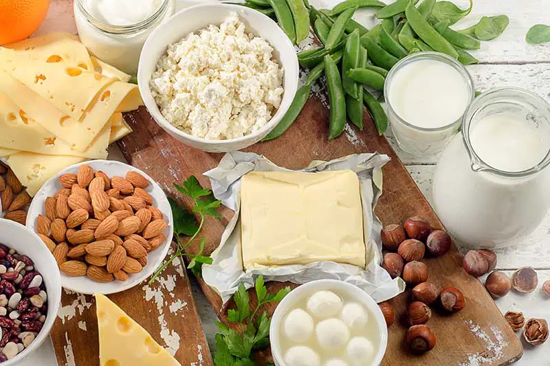 Una imagen horizontal de primer plano de una variedad de alimentos diferentes: frutos secos, queso, leche y verduras sobre una tabla de cortar de madera.