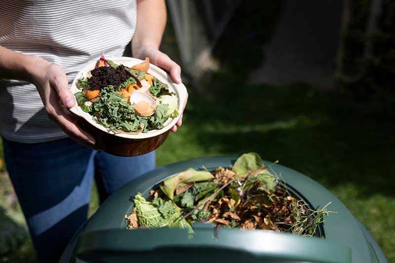 Una imagen horizontal de primer plano de un jardinero poniendo un cuenco lleno de restos de comida en un cubo de compostaje de plástico.