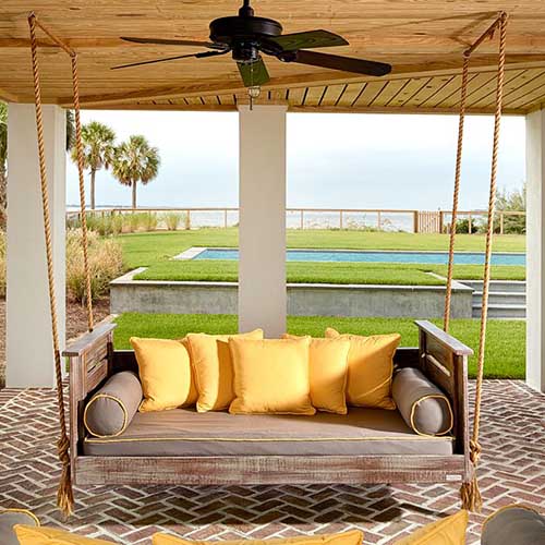 Un porche cubierto de lados abiertos con un banco de madera que cuelga del techo con vista a un jardín con una piscina y el océano al fondo.