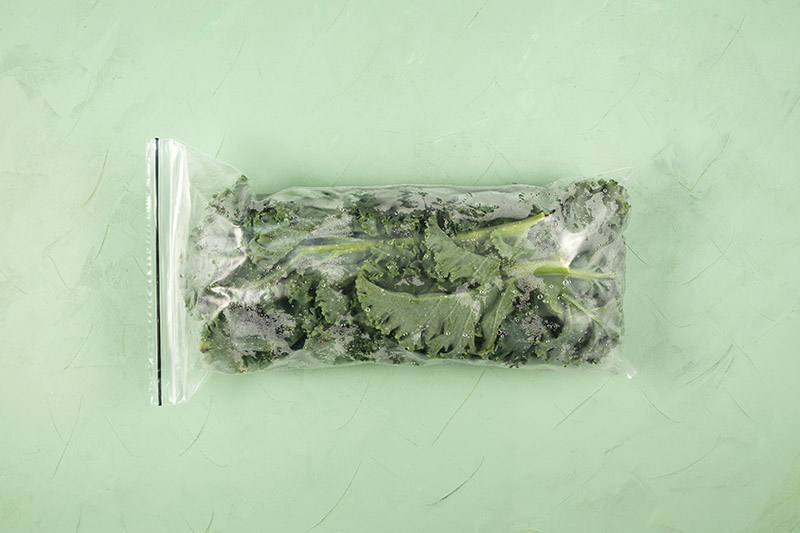 Una imagen horizontal de primer plano de una pequeña bolsa de plástico con verduras congeladas sobre una superficie verde.
