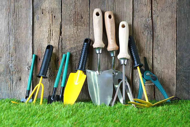 Una imagen horizontal de primer plano de un conjunto de herramientas de jardinería alineadas por una valla de madera.