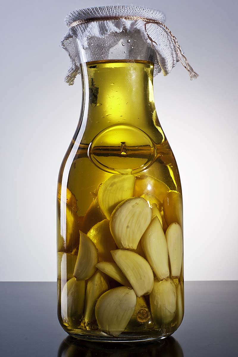 Una imagen vertical de cerca de una botella llena de aceite y ropa de ajo pelado sobre una superficie gris frente a un fondo blanco.