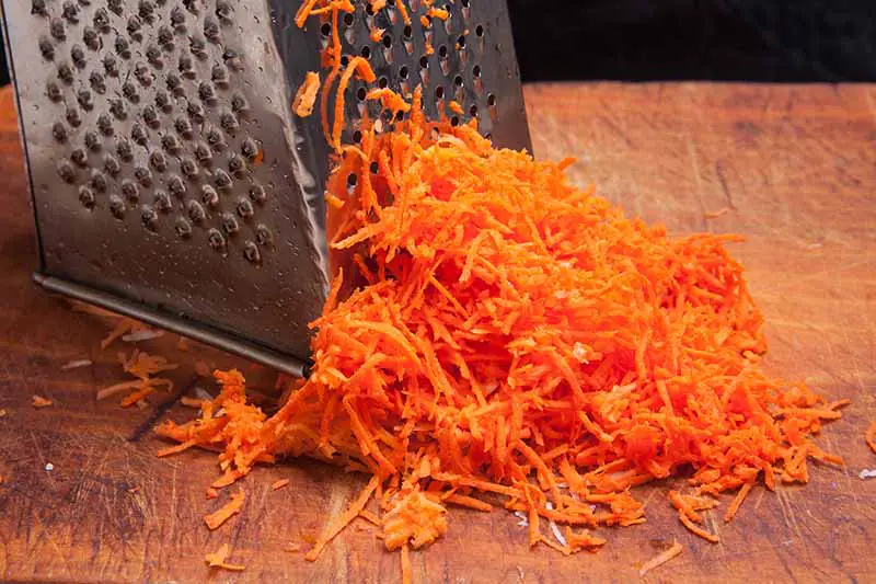 Un primer plano de un rallador de queso que se utiliza para rallar zanahorias en una tabla de cortar de madera sobre un fondo oscuro.