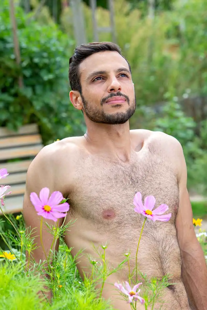 Una imagen vertical de un joven con el pecho desnudo sentado en el jardín con flores rosas en primer plano y un fondo de enfoque suave.