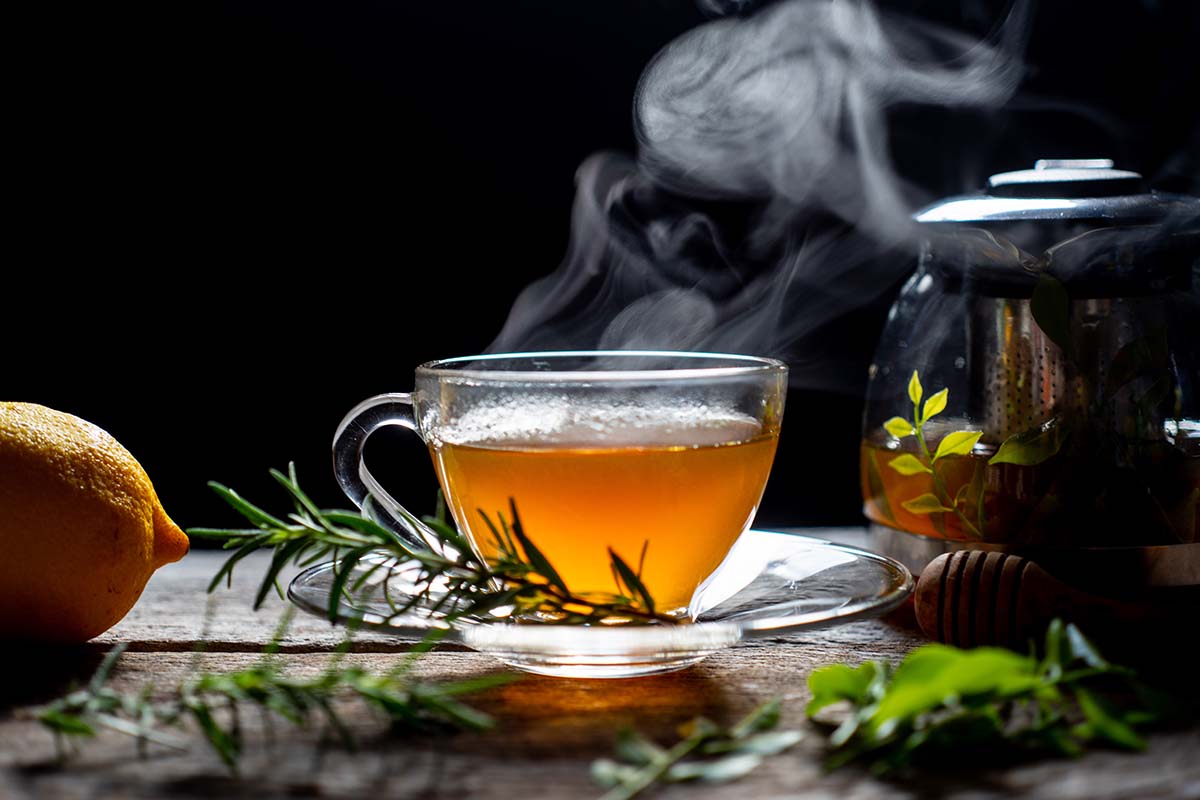 Una imagen horizontal de primer plano de té caliente en una taza de vidrio con vapor en un fondo oscuro.