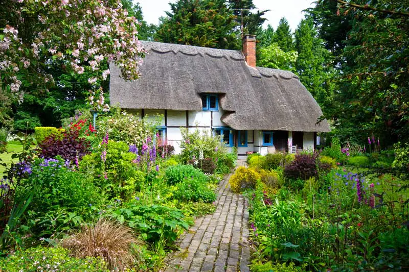 Jardín de cabaña con camino central que conduce a una casa Tudor de estilo rústico con techo de paja.