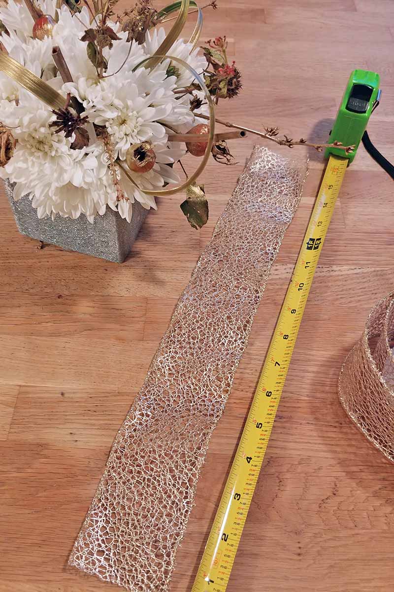 Se utiliza una cinta métrica amarilla con una caja de plástico verde para medir una cinta decorativa con alambre en una superficie de madera, con más cinta a la derecha y un arreglo floral a la izquierda.