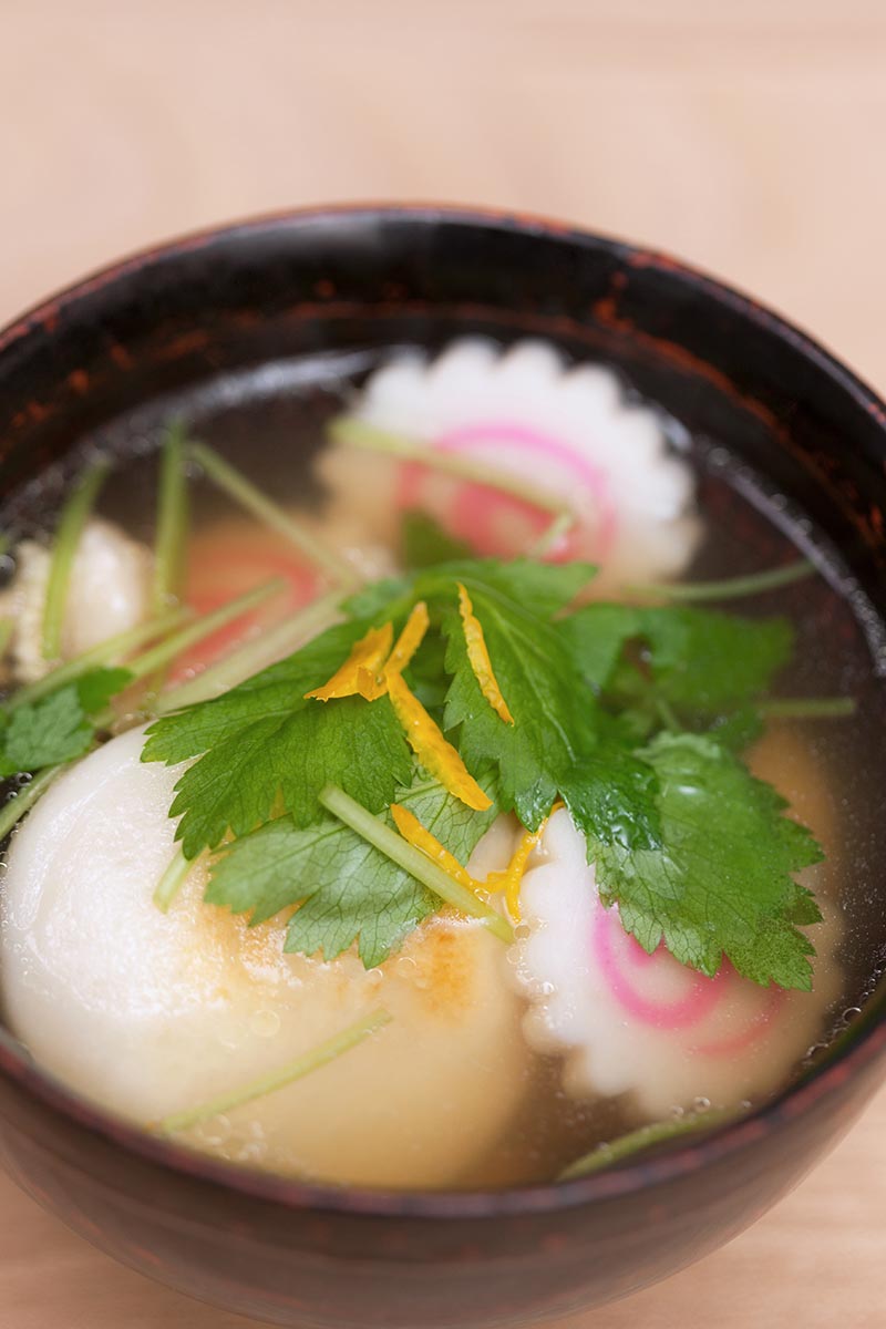 Una imagen vertical de cerca de una sopa clara con albóndigas adornadas con mitsuba en un fondo de enfoque suave.