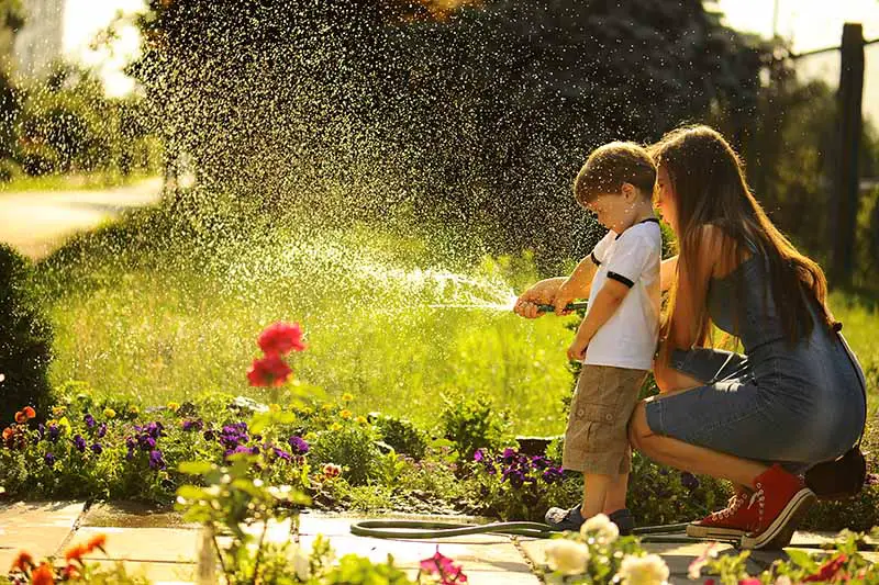 Una imagen horizontal de cerca de una madre y un niño regando el jardín con una manguera.