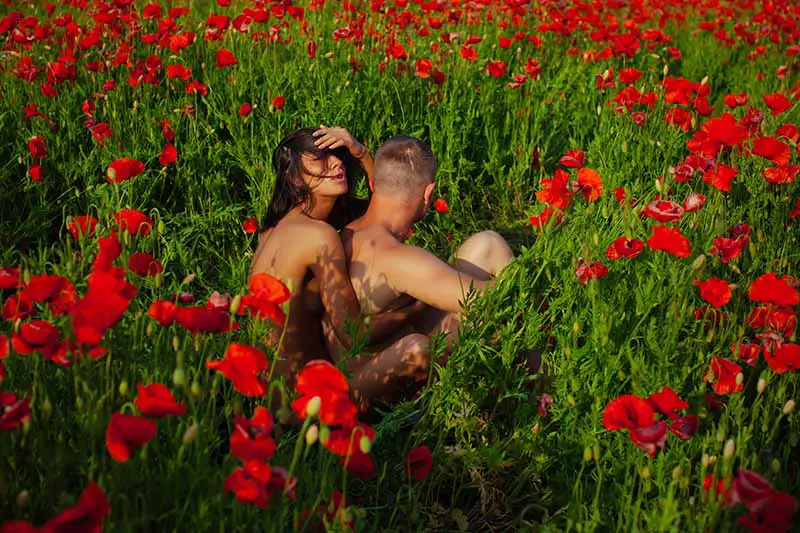 Un primer plano de un hombre con una mujer sentada detrás de él, desnudo en un campo de flores rojas y follaje verde suave bajo un sol brillante.