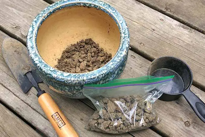 Una pequeña pala de mano descansa sobre una mesa de madera tosca junto a una vasija de cerámica y una bolsa que contienen muestras de tierra.  El suelo en ellos está seco y aglomerado.  A la derecha de todos ellos hay una pequeña taza de medir negra.