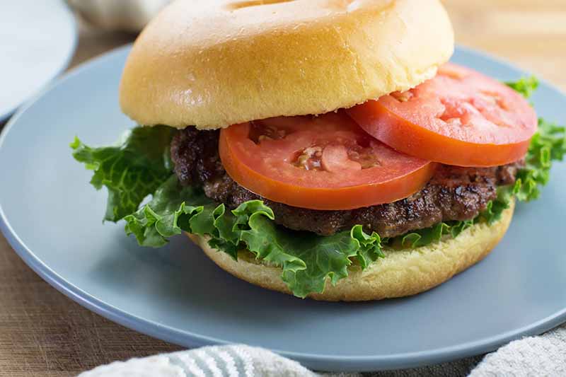 Una imagen horizontal de cerca de una hamburguesa casera con lechuga y tomate en un plato azul sobre una superficie de madera.
