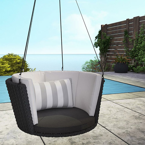 Un asiento colgante circular, en mimbre negro con cojines de rayas blancas, que cuelga sobre un patio de cemento con una piscina y el mar de fondo.
