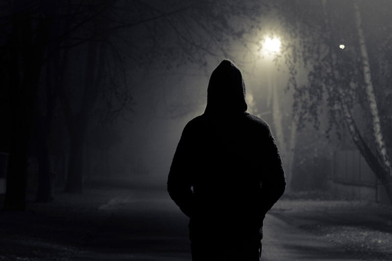 Una vista trasera de un hombre siniestro que lleva una capucha caminando por una calle en la noche con una farola para la luz.
