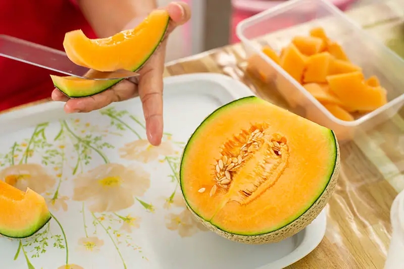 Un primer plano de una mano desde la izquierda del marco cortando un melón de color naranja brillante.  La otra mitad de la fruta se coloca en una bandeja de plástico sobre una superficie de madera.