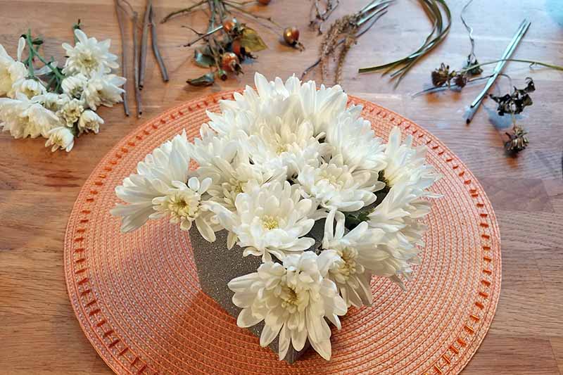 Crisantemos blancos en un recipiente cuadrado sobre un mantel individual redondo de color melón, sobre una superficie de madera marrón rodeada de flores cortadas y una variedad de vegetación de jardín.