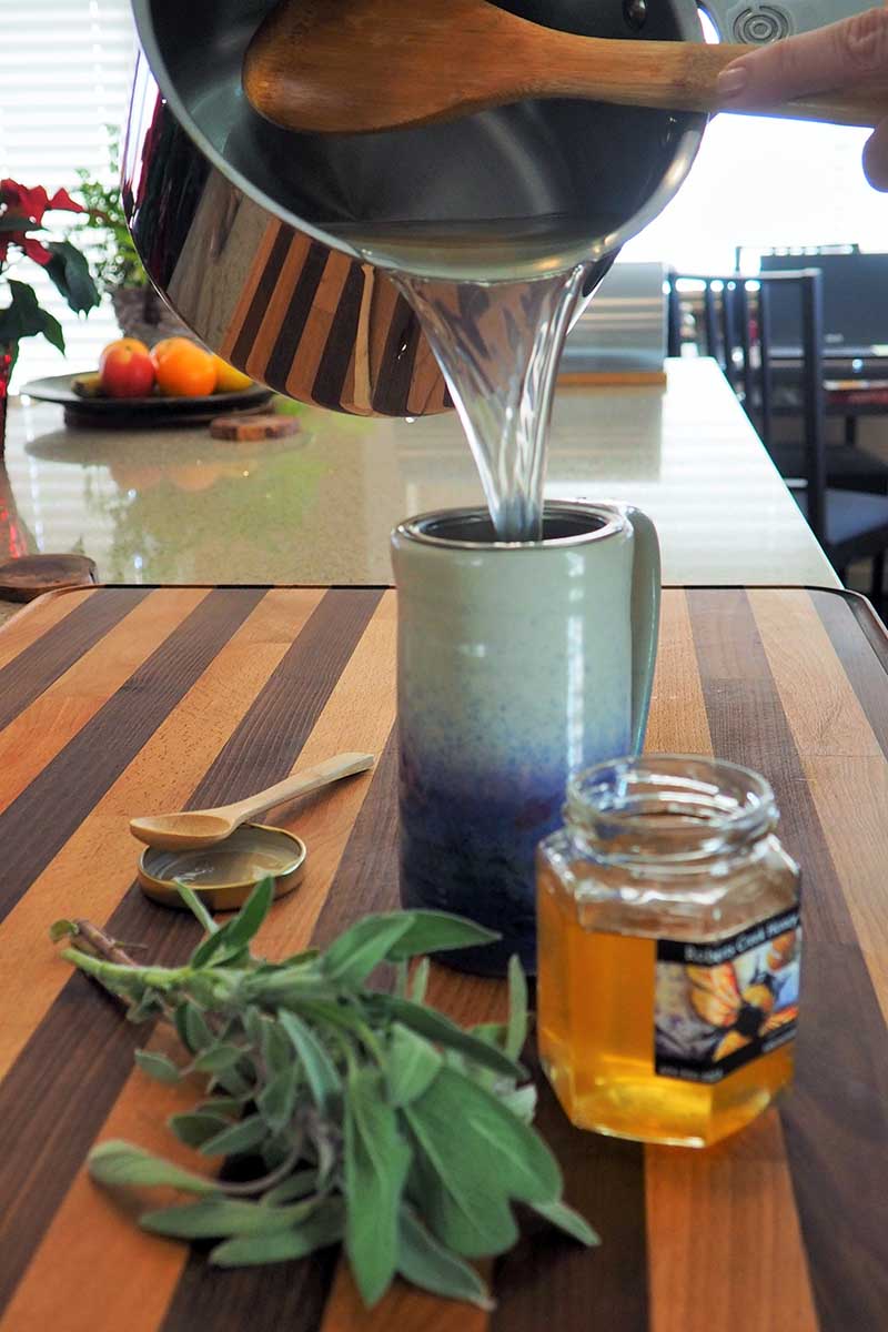 Una imagen vertical de primer plano de un té de hierbas que se vierte de una cacerola a una taza de cerámica sobre una superficie de madera.