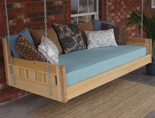 Un banco de madera con un colchón azul y coloridos cojines dispersos cuelga de cadenas en un porche con un edificio de ladrillo al fondo.