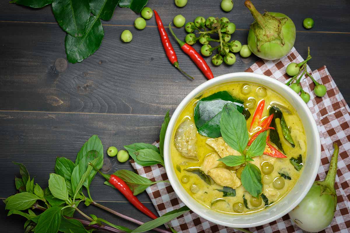 Una imagen horizontal de arriba hacia abajo de un curry tailandés recién preparado en un tazón blanco sobre un paño de cocina a cuadros sobre una superficie de madera, con verduras y hierbas esparcidas por todas partes.