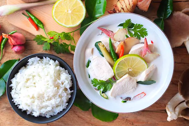 Una imagen horizontal de arriba hacia abajo de un tazón blanco con sopa de pollo tailandesa recién preparada sobre una superficie de madera con un tazón de arroz a la izquierda del marco y hierbas esparcidas alrededor.
