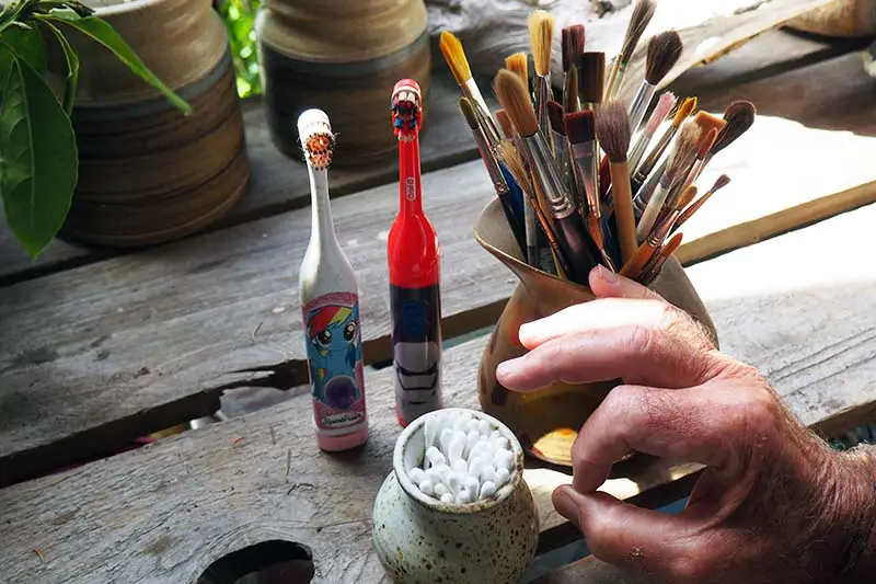 Una imagen horizontal de primer plano de una mesa de madera con dos cepillos de dientes que funcionan con pilas, un frasco lleno de pinceles de arte y un bote pequeño de bastoncillos de algodón, con una mano a la derecha del marco.