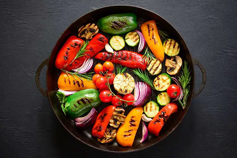 Una imagen horizontal de arriba hacia abajo de una sartén de hierro fundido con una selección de verduras asadas sobre una superficie gris oscuro.