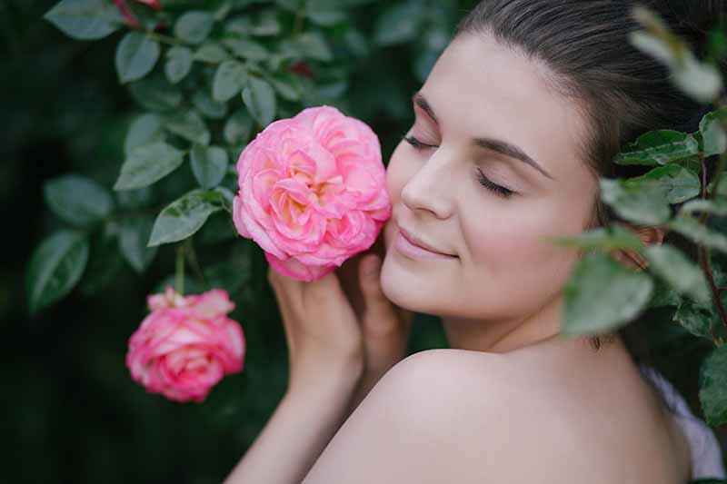 Un primer plano de una mujer joven con los hombros desnudos, ahuecando una flor rosa junto a su cara con los ojos cerrados, sobre un fondo de enfoque suave.