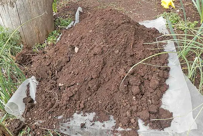 Una pila de tierra de color marrón oscuro con algunas rocas y raíces mezcladas, encima de una lona de plástico transparente en el jardín.