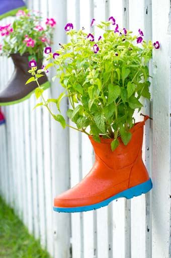 Dos botas de lluvia colgadas en una valla de estacas blancas, utilizadas como maceteros colgantes llenos de flores de espoleta rosadas y moradas.